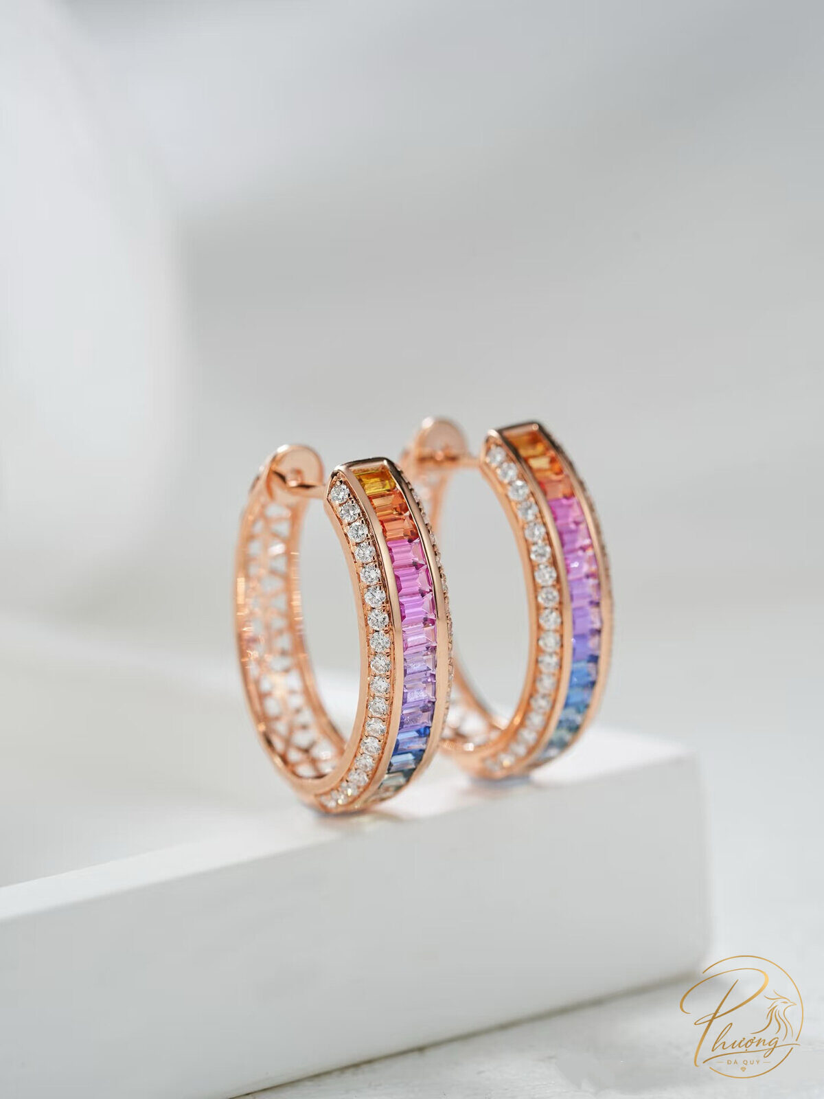 Khuyên tai vòng vàng hồng đính đá quý nhiều màu dáng ombre (cam, hồng, tím, xanh), đặt trên bệ trắng