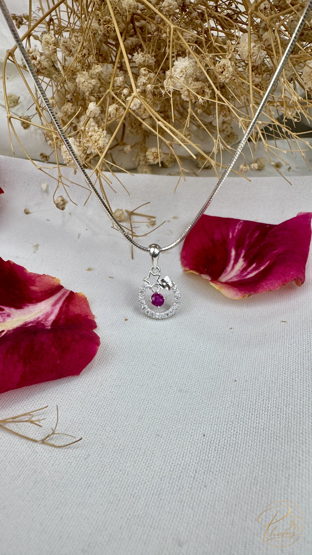 mặt dây chuyền bạc đính đá ruby nhỏ đặt cạnh hoa khô và cánh hoa hồng đỏ